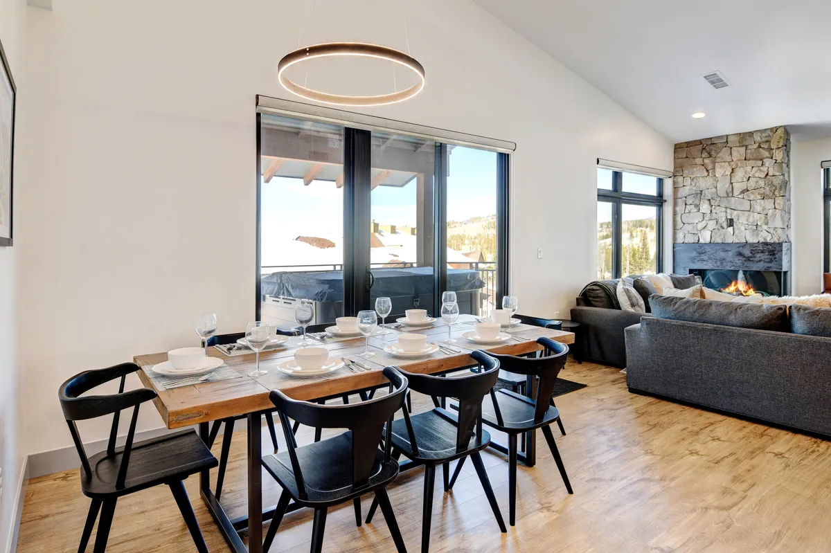 A dining table set for a meal adjacent to a comfortable seating area with a fireplace and large windows providing scenic views. The space is arranged to accommodate gatherings, celebrations, or quiet mornings appreciating the changing landscape outside.