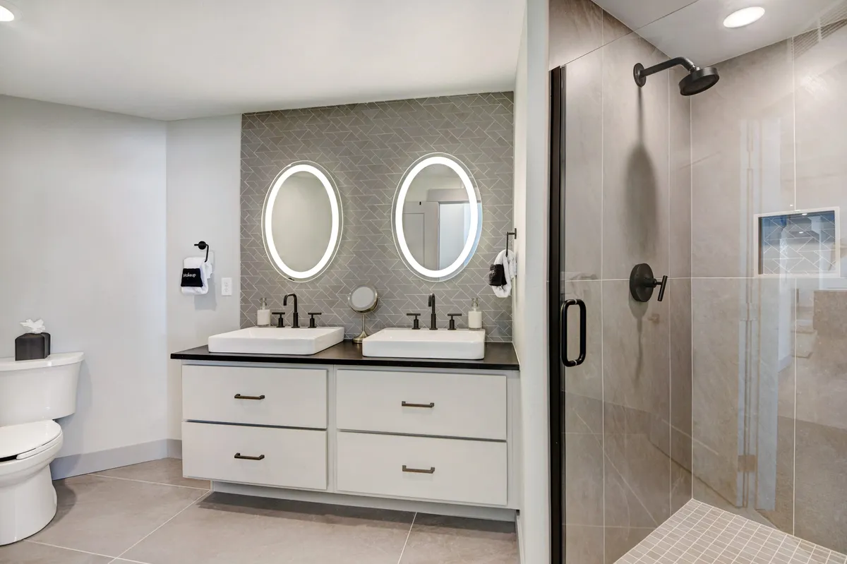 A modern ensuite bathroom featuring double vanity sinks with oval mirrors and a glass-enclosed walk-in shower. The space is bathed in soft natural light, emphasizing a refreshing, spa-like experience.