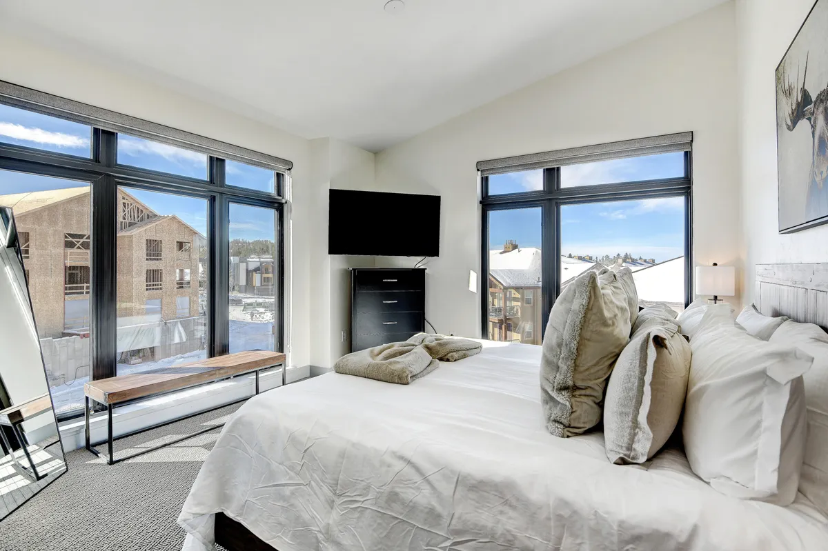 A serene bedroom with a large window, a bed featuring soft linens, and a TV mounted on the wall. The spacious window allows for views of the snowy landscape and abundant natural light, creating a restful retreat after a day of outdoor activities.