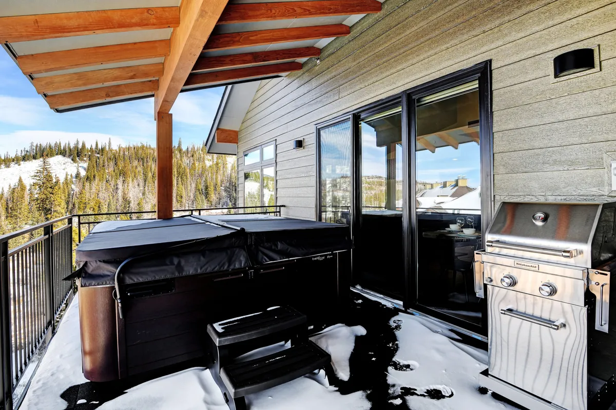 This exterior image features a private patio area with a hot tub and a BBQ grill, framed by wooden beams and overlooking scenic mountain views. The ambiance is tranquil and cozy, perfect for relaxing soaks and outdoor dining while surrounded by nature.