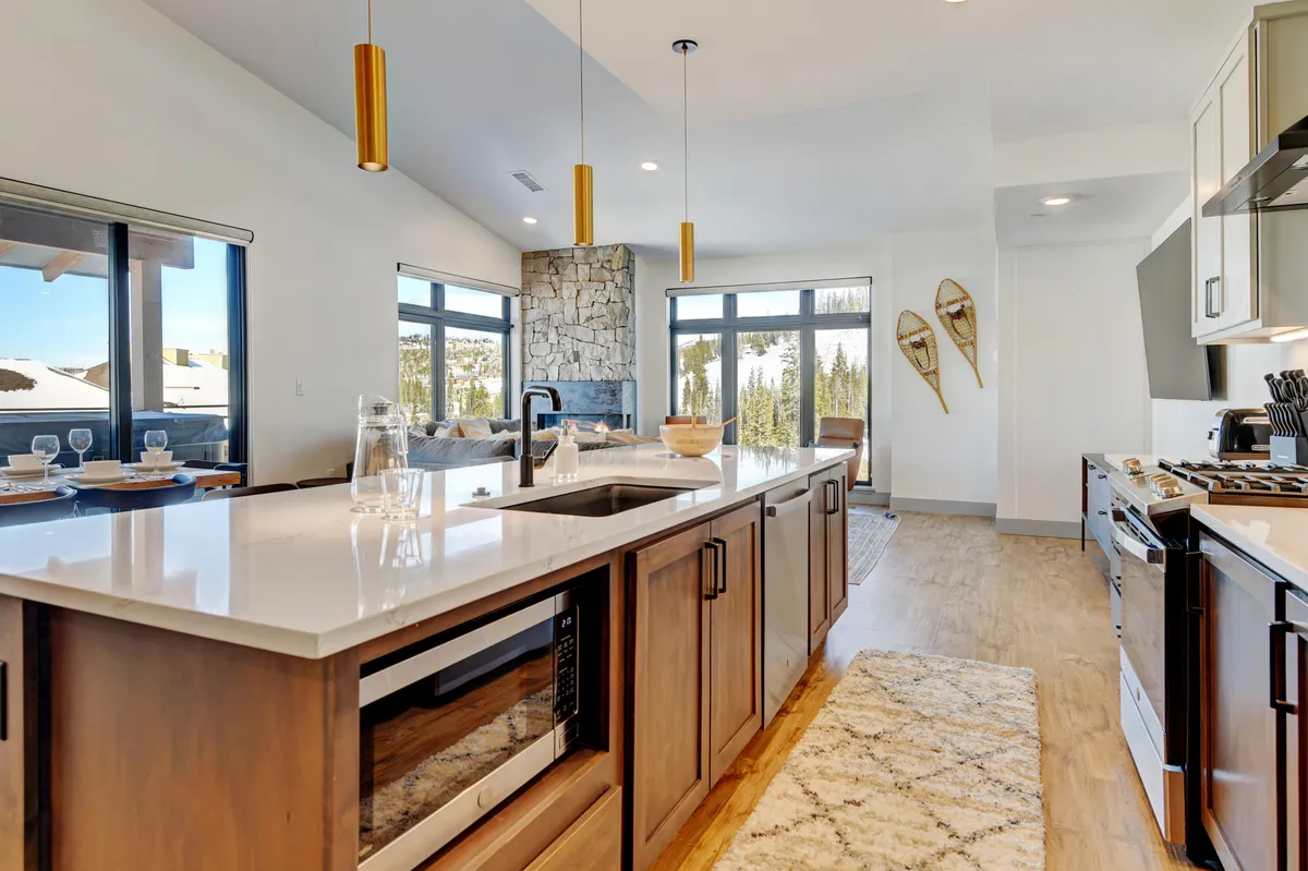 This image features a welcoming open-plan living space, with a prominent stone fireplace, expansive kitchen island, modern appliances, and large windows that fill the area with natural light. Decorative skis on the wall add a mountain touch, while the layout seamlessly connects the kitchen, dining, and living areas—perfect for gatherings.