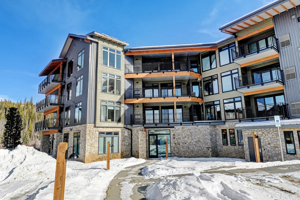 This exterior image features the Timber Fox building with its inviting stone facade, expansive windows, and wooden balconies. The wintry backdrop hints at the proximity to outdoor recreation while the balconies suggest spaces for private relaxation or social gatherings. The communal fire pit and snow-dusted setting reinforce the resort's atmosphere and its connection to the outdoors.