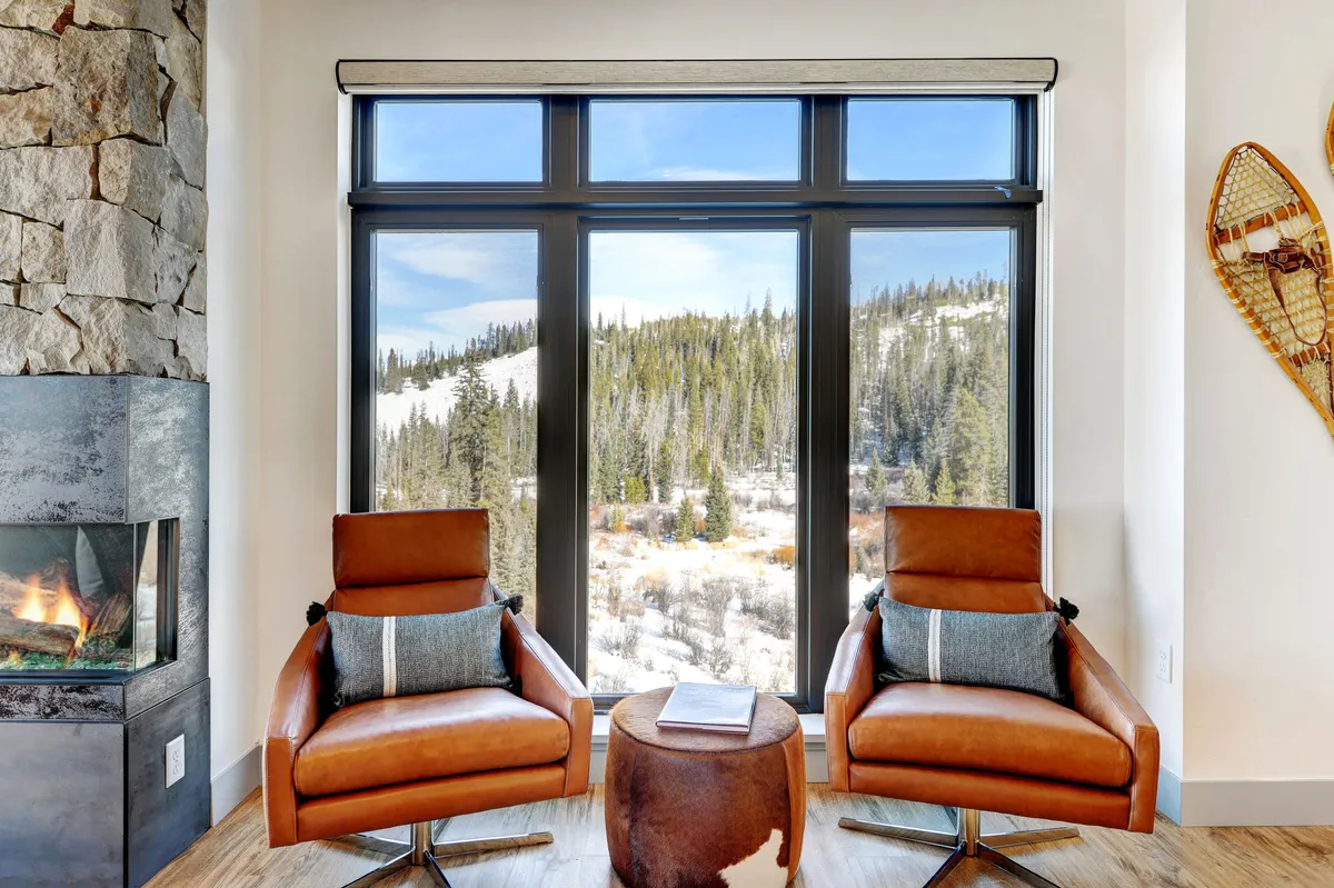 A cozy interior with large windows that frame a beautiful view of the mountains, brown leather chairs arranged for relaxation, a stone fireplace for warmth, and a wooden table. The outdoor scenery is visible, suggesting a strong connection between indoor comfort and the surrounding winter landscape.