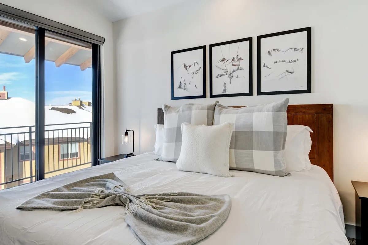 A king guestroom featuring a cozy bed with decorative pillows, framed artwork, and a door opening to a balcony with mountain views. Soft morning light fills the space, emphasizing both comfort and a connection to the outdoors.
