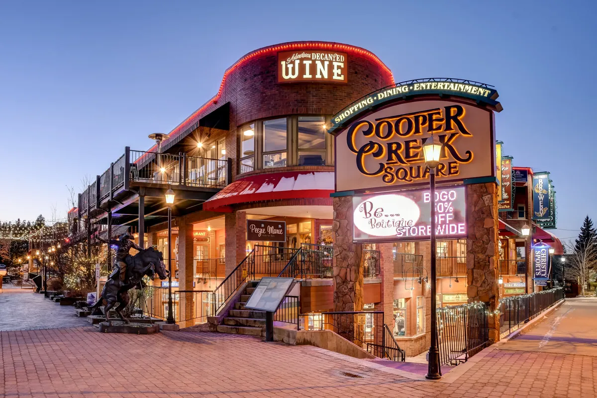 A vibrant plaza at Cooper Creek Square featuring a prominent sign, outdoor seating areas, illuminated shopfronts, and a striking horse sculpture. People gather around, hinting at a lively mix of dining, shopping, and socializing opportunities.