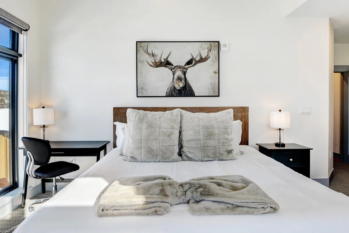 A spacious bedroom centered around a large plush bed adorned with decorative pillows, flanked by nightstands and lamps, with a moose artwork above the headboard. A private desk and chair are positioned by the bed, ideal for remote work. Natural light pours in through a nearby window, creating a serene and restful atmosphere.