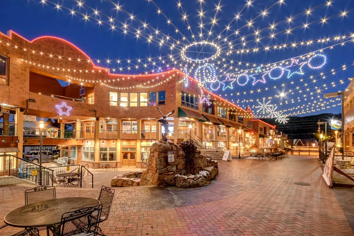 An inviting exterior scene featuring twinkling string lights overhead, a decorative stone fountain, and boutique shops with warmly illuminated windows. People are mingling, enjoying al fresco dining, and the atmosphere hints at a lively community hub just steps from the property.