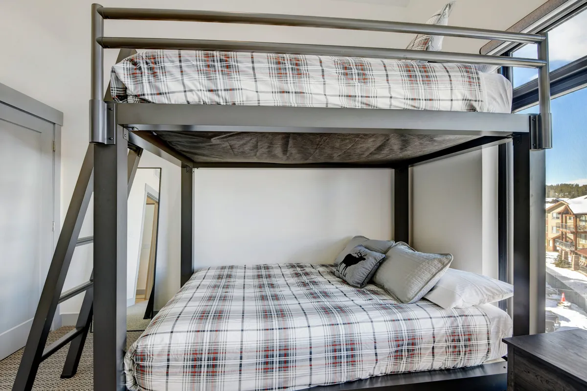 A cheerful bunk room designed for kids or friends, with a queen-over-queen bunk bed dressed in playful plaid bedding, a large window letting in daylight, and ample space for activities and relaxation.