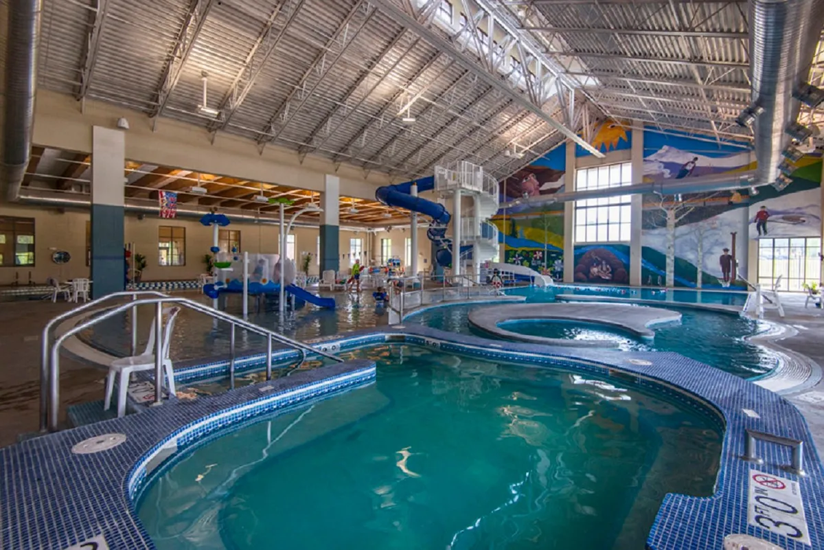 A family-friendly recreation center interior featuring a large swimming pool, colorful water slides, playful murals, and dedicated play areas for children. The atmosphere is energetic and welcoming for all ages.