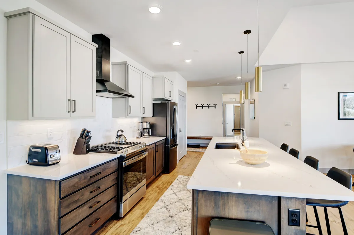 A modern, functional kitchen boasting a large central island with bar seating, stainless steel appliances, and warm wooden cabinetry. The inviting layout is perfect for cooking, entertaining, and sharing meals, illuminated by elegant lighting fixtures overhead.